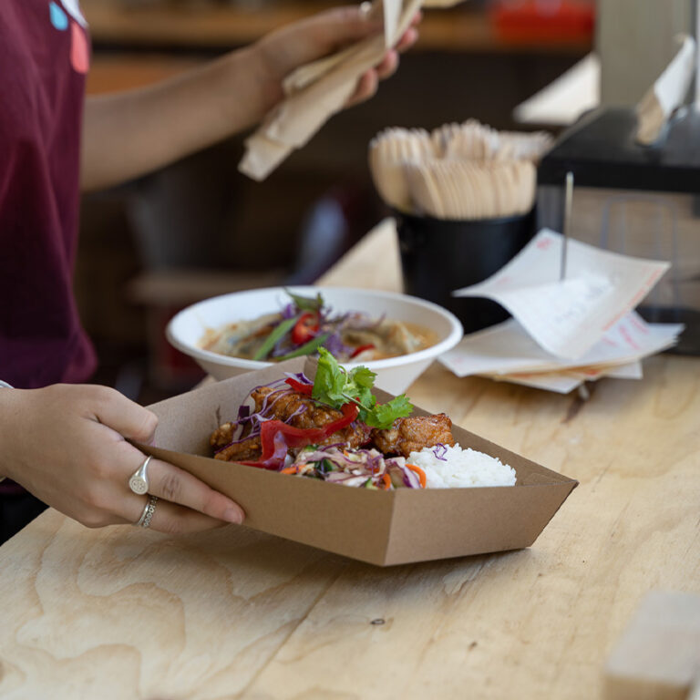 Asian takeaway food in a compostable BioBoard container. Soup in a plant fibre bowl in the background, and the person serving the food is holding napkins and wooden cutlery suitable for composting.